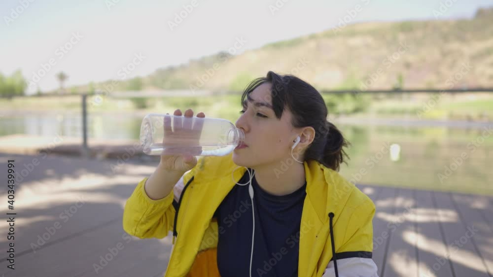 Fitness woman drinks water from her bottle while listening to music and resting in slow motion Fitness woman drinks water from her bottle while listening to music and resting in slow motion