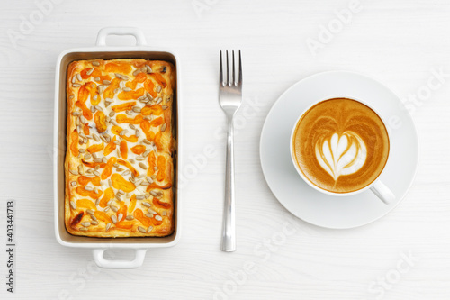 Cup of coffee and homemade cottage cheese casserole with dried apricots and sunflower seeds on white wooden table. Top view.
