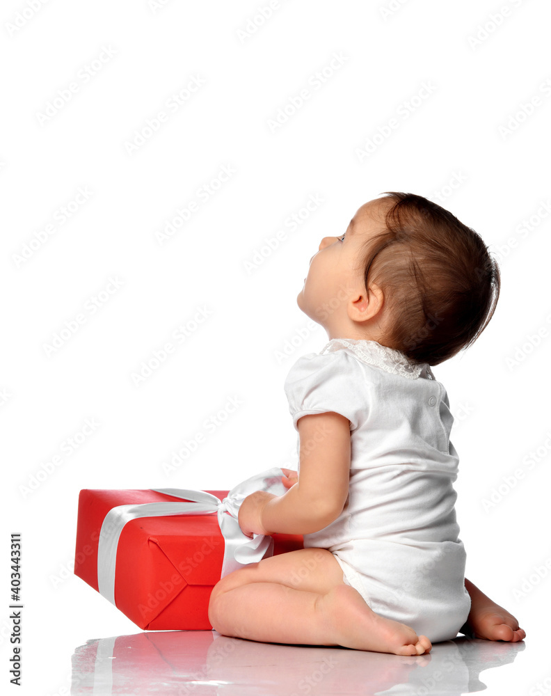 Baby and wrapped holiday gift boxes with bow. Surprised infant girl wearing bodysuit, sitting on white studio floor background, looking to the side. Isolated portrait. Birthday or Christmas concept Baby and wrapped holiday gift boxes with bow. Surprised infant girl wearing bodysuit, sitting on white studio floor background, looking to the side. Isolated portrait. Birthday or Christmas concept