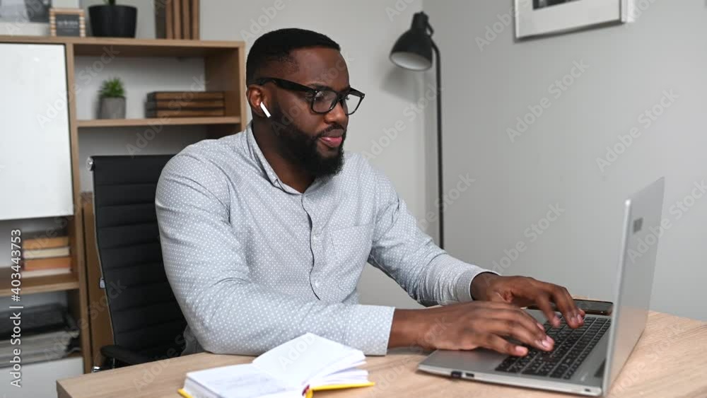 Positive African-American guy in glasses is using laptop for remote work from home. A multiracial young man is typing message or answering email sitting at the desk