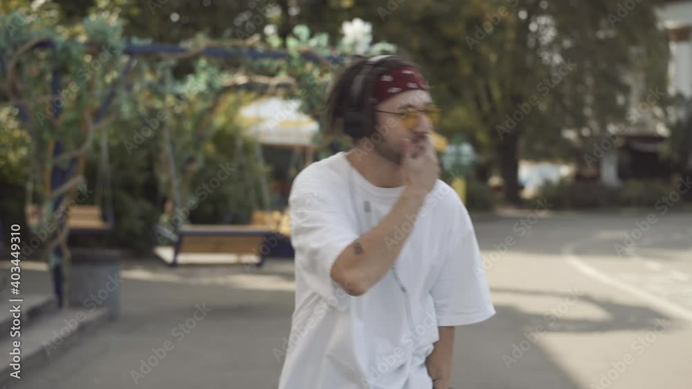Relaxed tattooed Caucasian young man with dreadlocks walking along sunny urban street dancing to music in headphones. Portrait of carefree millennial guy enjoying leisure outdoors.