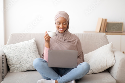 Joyful black woman in hijab having video chat with family or friend on laptop at home, drinking coffee on cozy couch