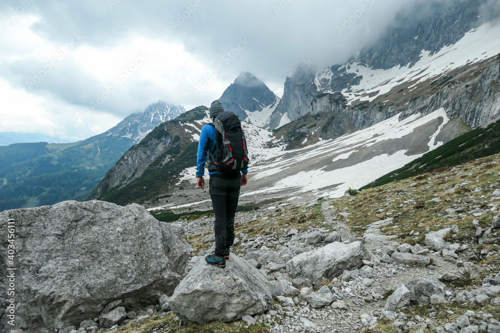 A backpacker man in a beanie hiking in high Alps in the region of Dachstein, Austria. He is crossing a landslide area. He is walking alone, enjoying the view on the vast valley. Overcast. Solitude