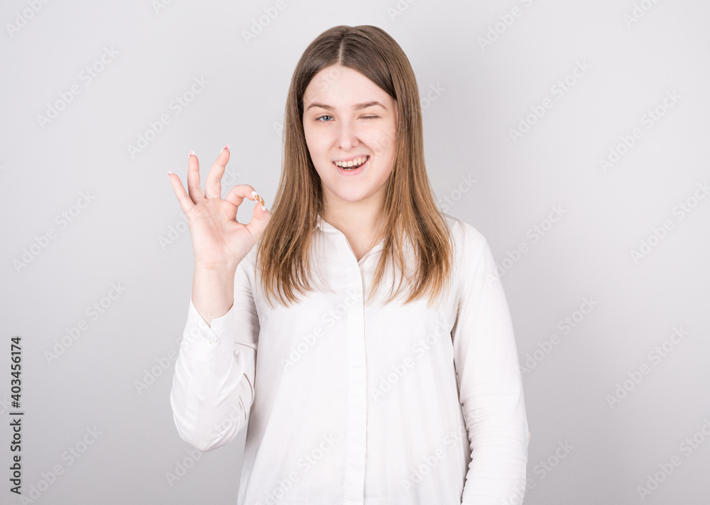 Close-up photo of funny young brunette woman in white shirt showing OK gesture, looking at camera, isolated on gray background Close-up photo of funny young brunette woman in white shirt showing OK gesture, looking at camera, isolated on gray background