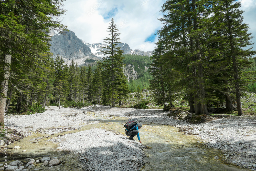 A backpacker man drinking from a rushing torrent in region of Dachstein, Austria. There is dense forest behind, and high Alpine peaks. Taking a moment to relax. Admiring the beauty of the nature.