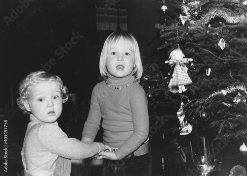 December 1977 vintage, retro monochrome image of a brother and sister, holding hands, standing next to a vintage decorated christmas tree. 