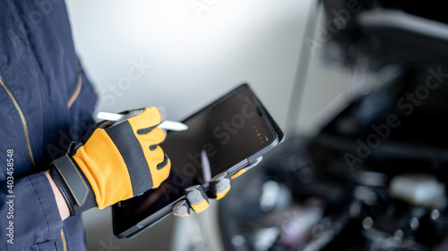 Male auto mechanic with protective suit using digital tablet for checking car in auto service garage. Mechanical maintenance engineer working in automotive industry. Automobile servicing and repair