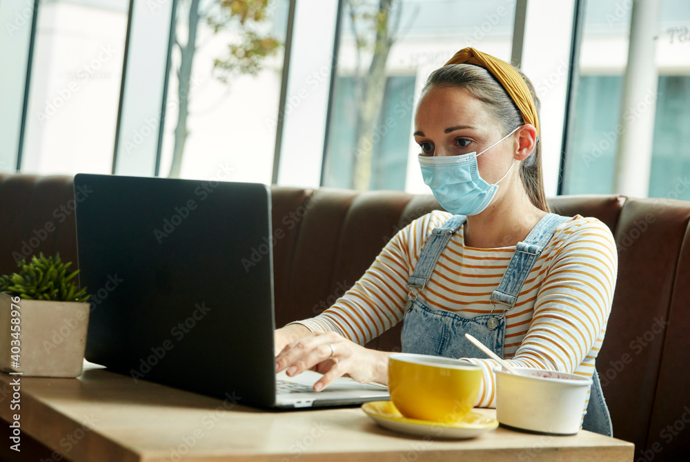 Woman wearing a face mask seated in a cafe using a laptop Woman wearing a face mask seated in a cafe using a laptop