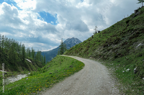 A gravelled road through Alpine valley in Austria in the region of Dachstein. High mountain around, partially covered with snow. Stony and sharp mountains. Overcast. Dense forest at the foothill. photo