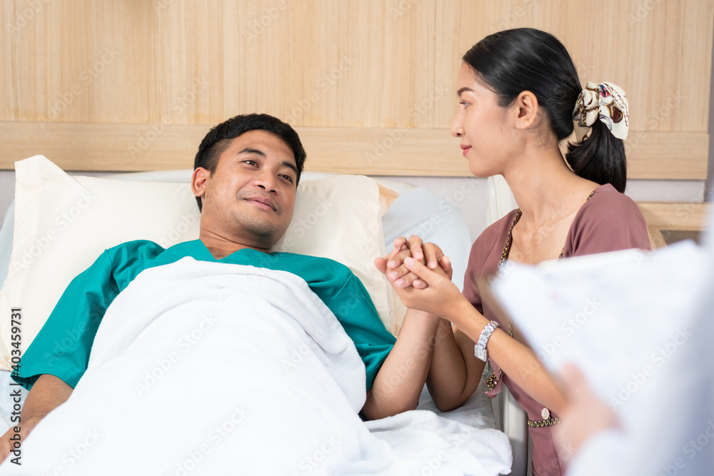 Happy Asian wife holding husband hand and look at him with smile. patient man lying in bed health getting well and stronger. support woman and take care at hospital concept Happy Asian wife holding husband hand and look at him with smile. patient man lying in bed health getting well and stronger. support woman and take care at hospital concept