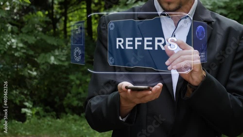 Unrecognizable businessman activates conceptual HUD holograms on smartphone with text Refers. Bearded man in white shirt and acket with holographic screen on a background of green trees photo