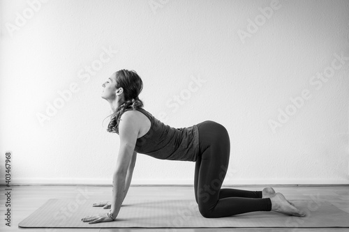 Side view of a young woman performing the Cow Pose or Bitilasana yoga pose in her home interior.