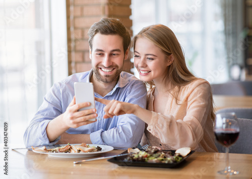 Cheerful millennial couple browsing internet on smartphone during romantic dinner at cozy cafe