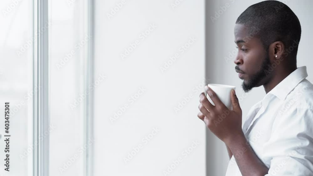Office anxiety. Coffee break. Work stress. Creative block. Disturbed worried African man employee in white shirt thinking about project deadline with hot drink at window. Office anxiety. Coffee break. Work stress. Creative block. Disturbed worried African man employee in white shirt thinking about project deadline with hot drink at window.
