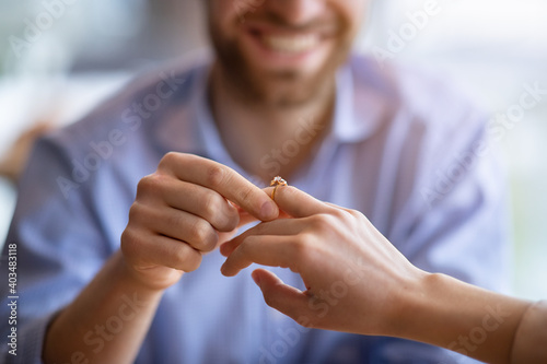 Happy guy holding his fiancee s hand  putting on engagement ring on her finger  closeup view