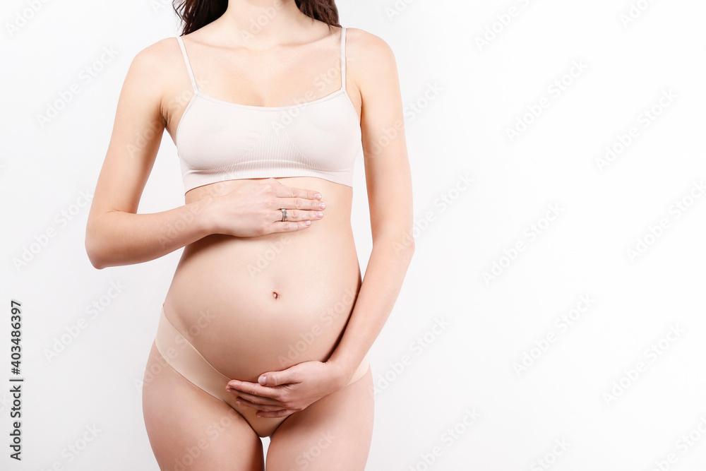 Pregnant woman wearing supportive seamless maternity bra and body color bottoms, arms on her belly. Female hands wrapped around bare tummy. Pregnancy underwear concept. Background, copy space. Pregnant woman wearing supportive seamless maternity bra and body color bottoms, arms on her belly. Female hands wrapped around bare tummy. Pregnancy underwear concept. Background, copy space.