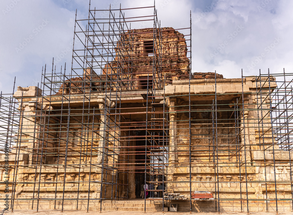 Hampi, Karnataka, India - November 5, 2013: Sri Krishna temple in ruins. Yellow-brown stone entry gate with gopuram under repair fronted by metal scaffold under blue cloudscape.