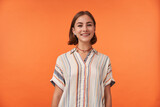 Portrait of girl with brown short haircut and braces for teeth, pierced nose, wearing striped shirt. Young smiling girl watching camera against orange background