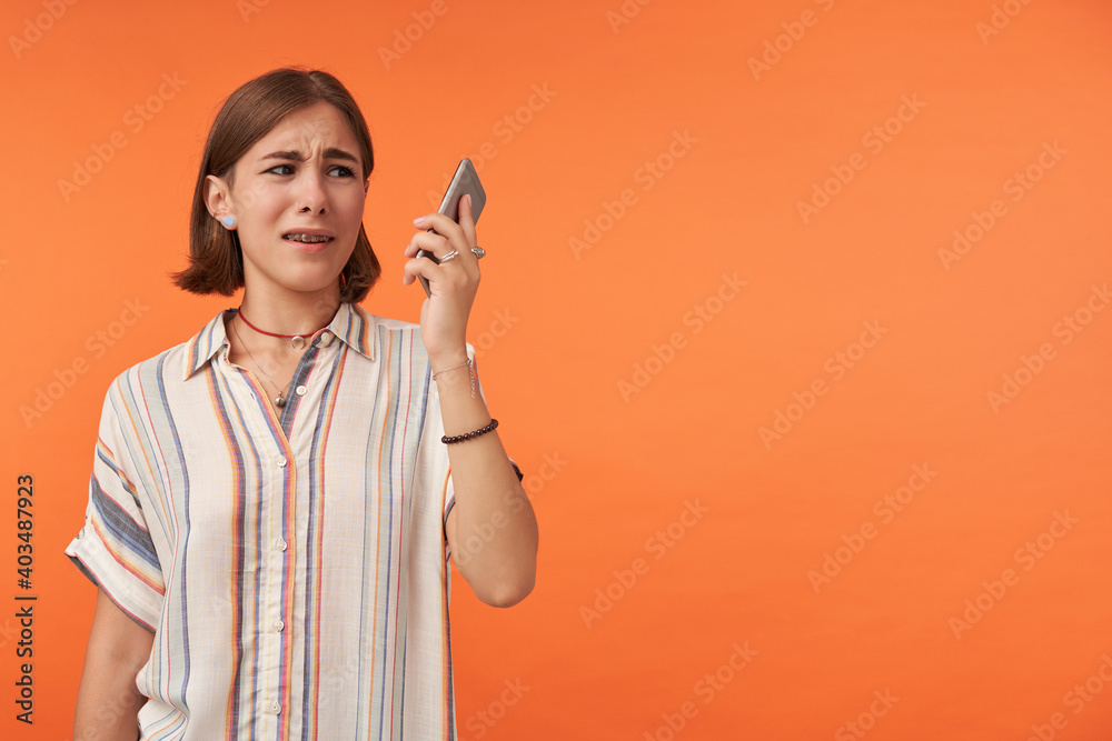 Unhappy girl hold and look to the smartphone. Did not like what she hear and shows it. Wearing striped shirt, teeth braces and bracelets. Watching to the right, copy space over orange background Unhappy girl hold and look to the smartphone. Did not like what she hear and shows it. Wearing striped shirt, teeth braces and bracelets. Watching to the right, copy space over orange background