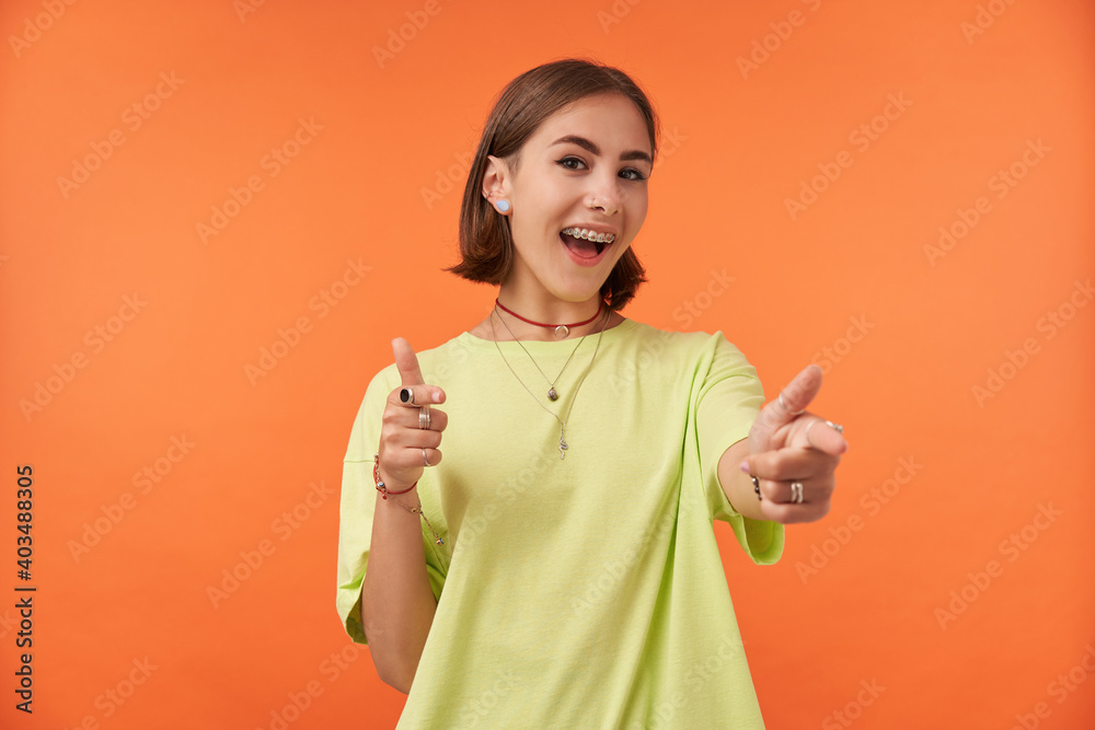 Portrait of nice looking, cheerful girl pointing to the camera and have wide smile. Happy looking. Wearing green t-shirt, teeth braces, bracelets and rings. Isolated against orange background Portrait of nice looking, cheerful girl pointing to the camera and have wide smile. Happy looking. Wearing green t-shirt, teeth braces, bracelets and rings. Isolated against orange background
