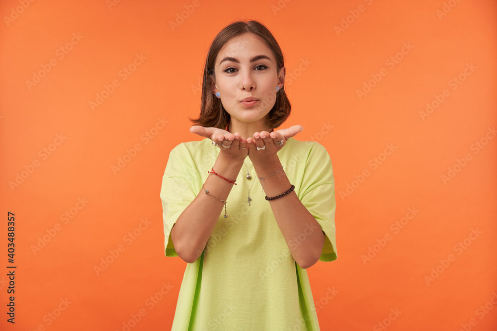 Portrait of attractive, nice looking girl with short brunette hair, sending a kiss, tempting. Look to the camera over orange background. Wearing green t-shirt, bracelets and rings Portrait of attractive, nice looking girl with short brunette hair, sending a kiss, tempting. Look to the camera over orange background. Wearing green t-shirt, bracelets and rings