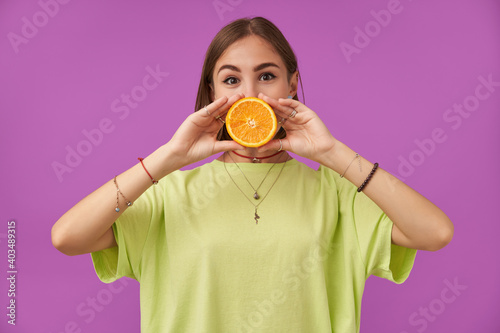 Portrait of attractive  nice looking girl holding orange over her mouth with two hands. Standing over purple background. Wearing green t-shirt  bracelets rings and necklace