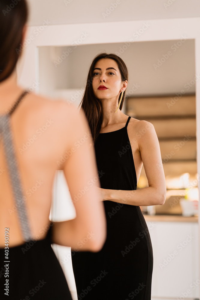 Woman in a long black dress with bare back looking at her reflection in the mirror while getting ready for an event, party, date Woman in a long black dress with bare back looking at her reflection in the mirror while getting ready for an event, party, date