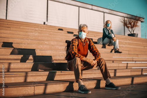 Senior man sitting on stairs and wearing protective surgical face mask. In background sitting senior woman wearing mask, too. Seniors esteem social distance during corona virus pandemic.