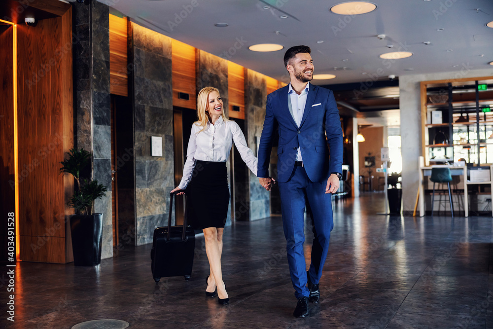 Smiling middle-aged couple holding hands and walking in the lobby of the fancy hotel. They going into their room. The woman is pulling a suitcase.
