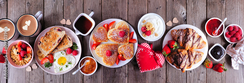 Valentines or Mothers Day brunch table scene. Overhead view on a dark wood banner background. Heart shaped pancakes, eggs and a variety of love themed food.