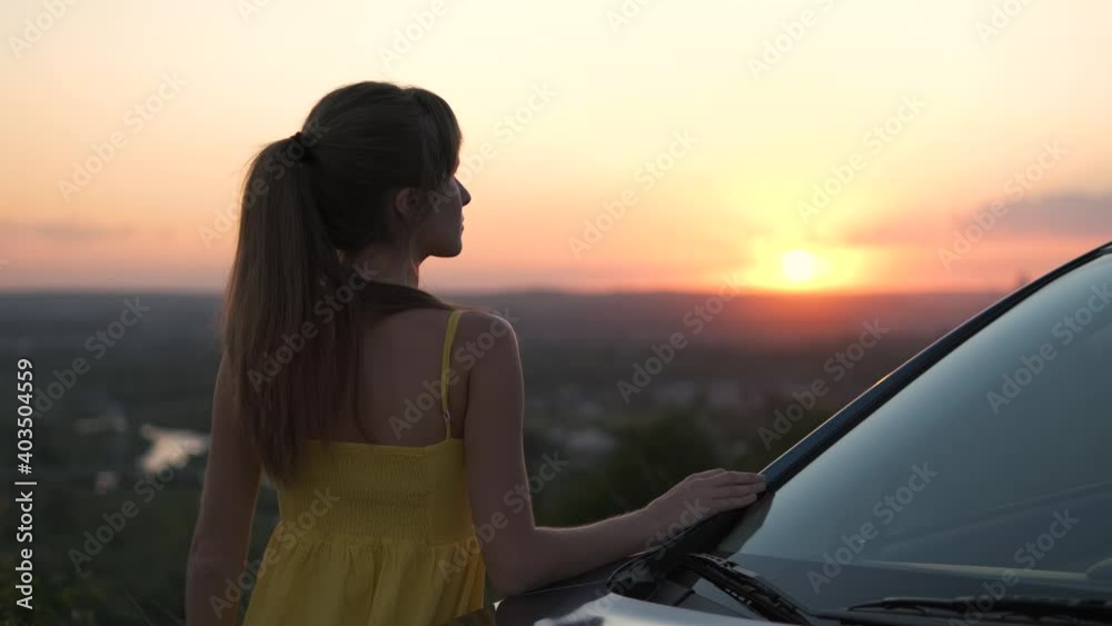 Young woman in summer dress standing near her car enjoying sunset view. Travel and vacations concept. Young woman in summer dress standing near her car enjoying sunset view. Travel and vacations concept.