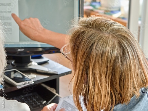 A woman, only recognizable from behind with her head, points with her right, outstretched arm at the screen of a PC to explain something.