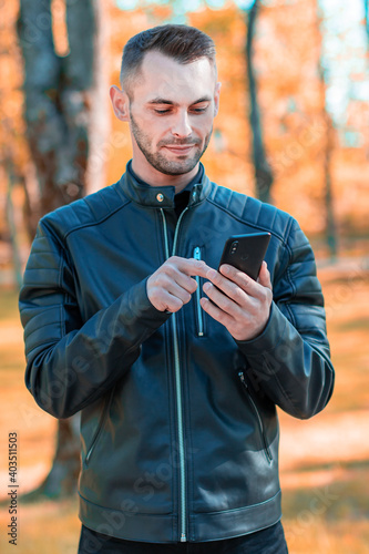 Fototapeta Naklejka Na Ścianę i Meble -  Youthful Guy Using Black Smartphone at the Beautiful Autumn Park. Handsome Young Man with Mobile Phone at Sunny Day - Medium Shot Portrait