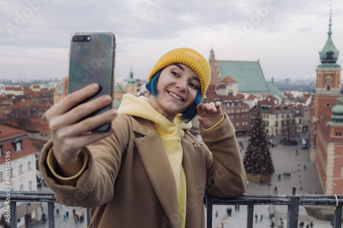 Happy young woman with colored blue hair in white coat taking selfie in front of old town and Christmas tree photo