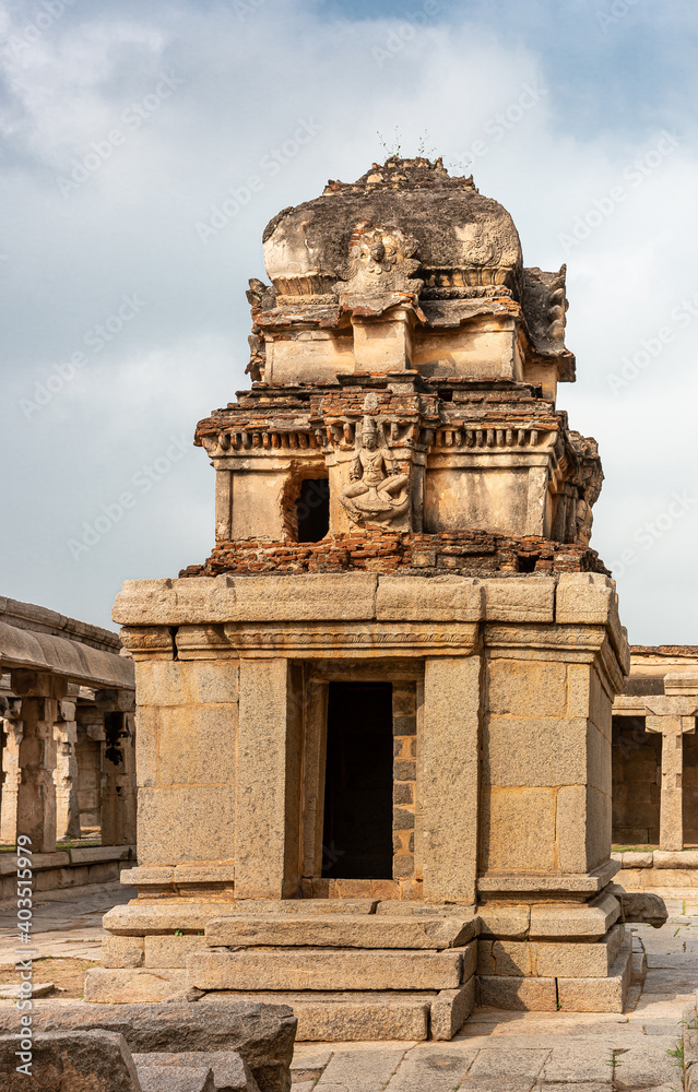 Hampi, Karnataka, India - November 5, 2013: Sri Krishna temple in ruins. Brown stone damaged small shrine with molded vimanam partly restored with red bricks under blue cloudscape.