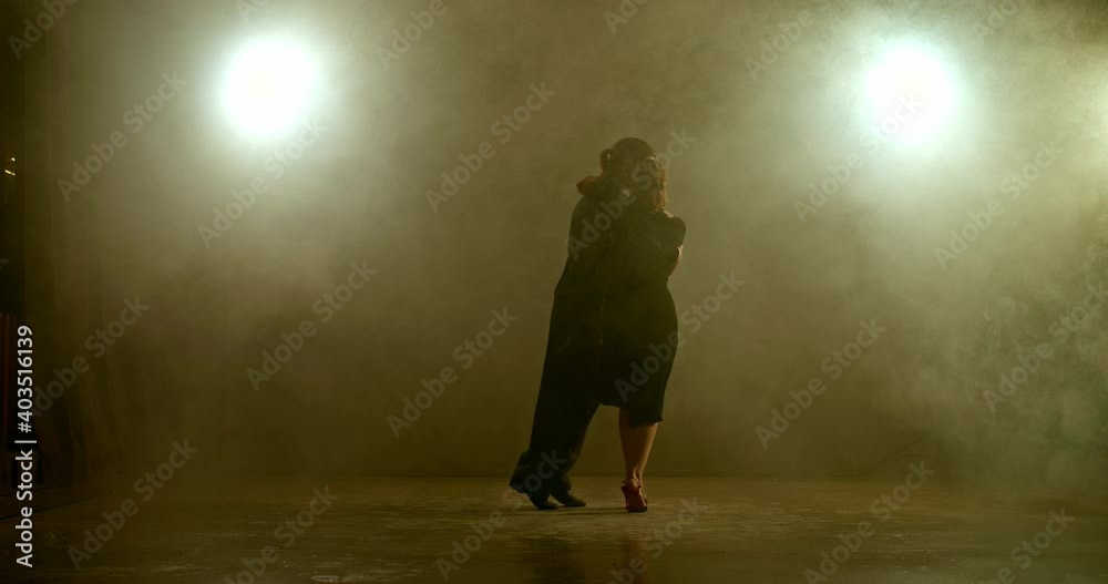 Beautiful romantic couple is dancing in a dark room with smoke, yellow lamps are shining. Elegant man in a black suit is dancing a beautiful woman, professional tango dancers. 4k, ProRes Beautiful romantic couple is dancing in a dark room with smoke, yellow lamps are shining. Elegant man in a black suit is dancing a beautiful woman, professional tango dancers. 4k, ProRes