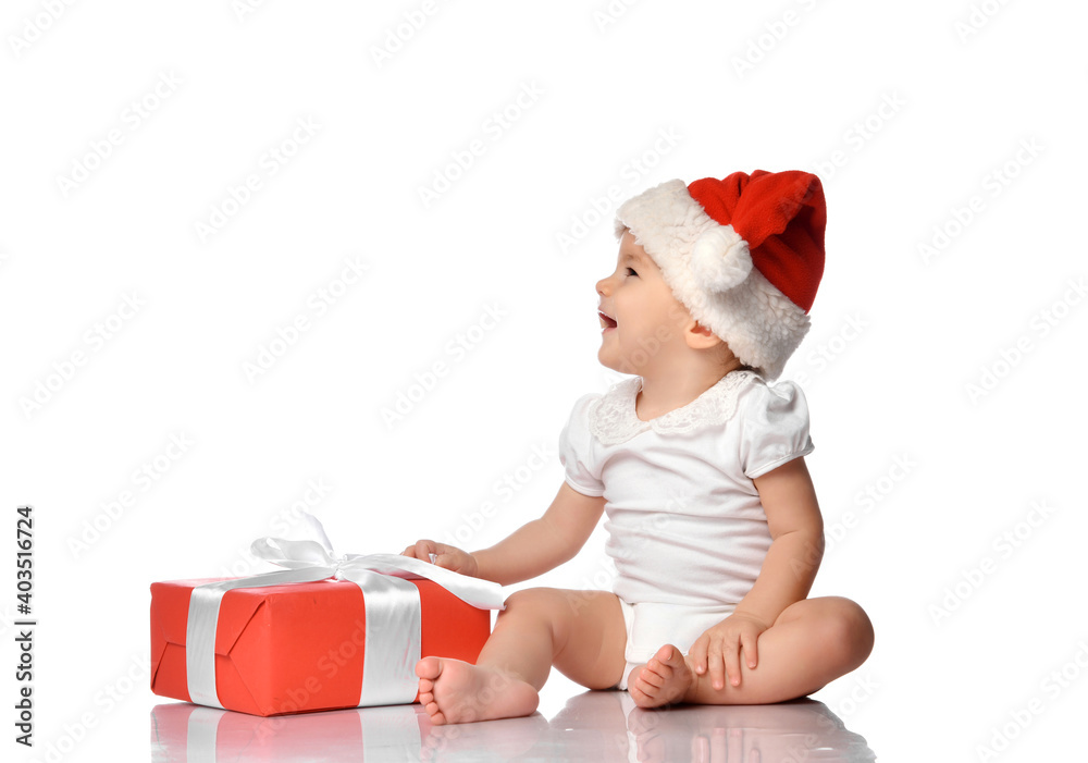 Cute laughing baby girl child wearing jumpsuit, red Santa Claus hat sitting with wrapped festive gift box on white floor studio background looking aside. Isolated portrait. Christmas, New Year concept Cute laughing baby girl child wearing jumpsuit, red Santa Claus hat sitting with wrapped festive gift box on white floor studio background looking aside. Isolated portrait. Christmas, New Year concept