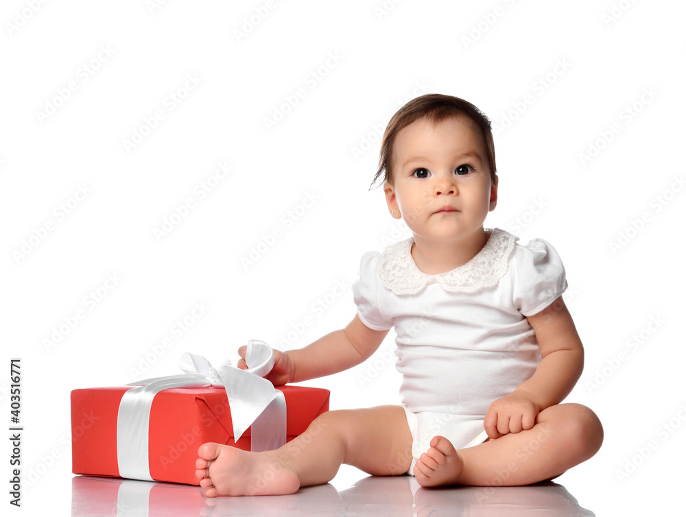 Happy little toddler sits on the floor in a white studio next to a gift wrapped in red wrapping paper. expectation of surprise and celebration Happy little toddler sits on the floor in a white studio next to a gift wrapped in red wrapping paper. expectation of surprise and celebration