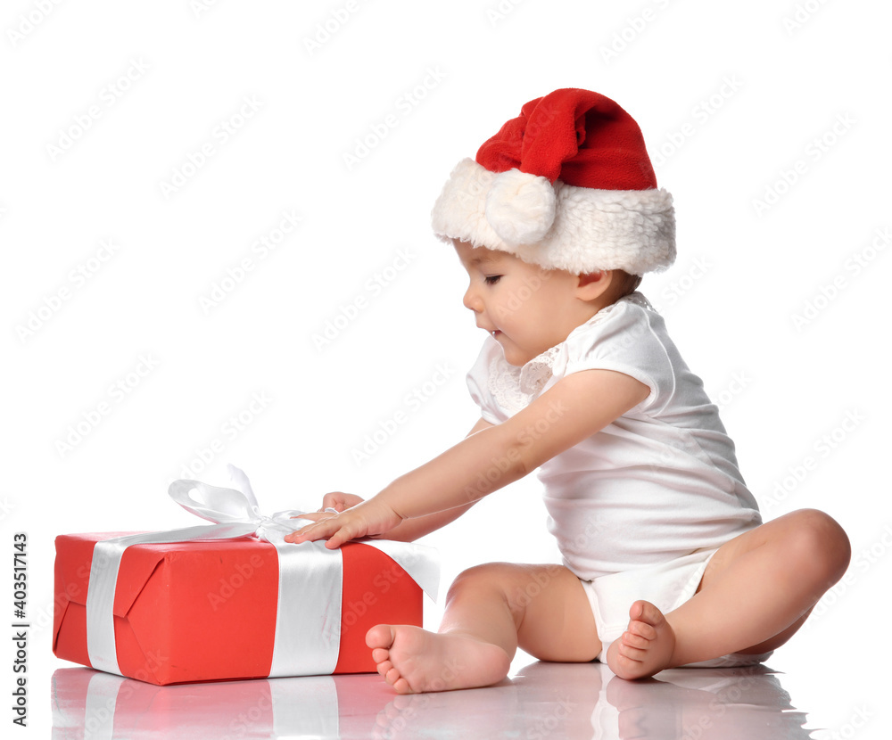 Cute infant baby girl wearing overalls, a red Santa Claus hat, sitting on a white floor studio, looking at a festive gift box wrapped with a bow. Isolated portrait. Christmas, New Year concept