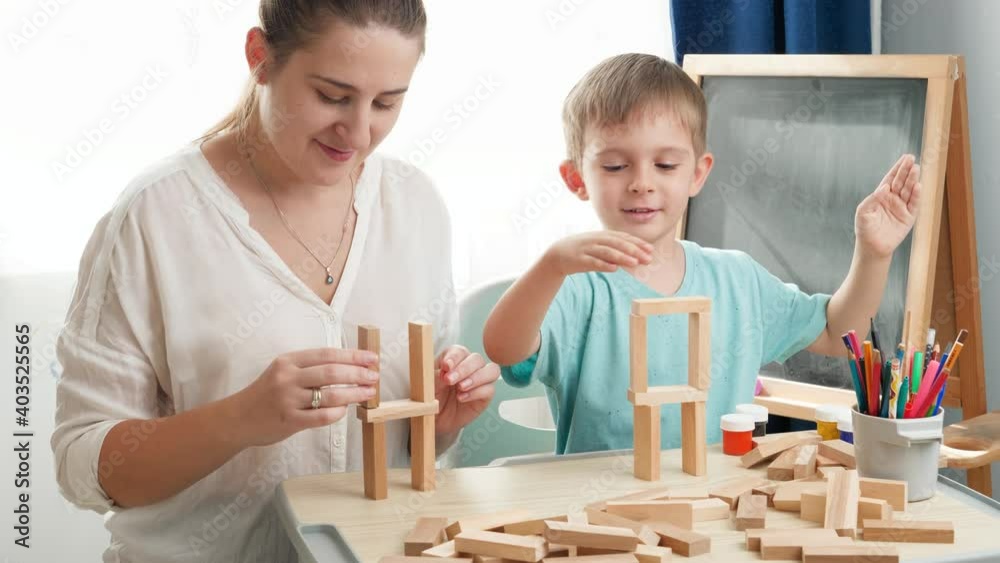 Slow motion video of young mother with little son playing and building high tower with wooden blocks or bricks. Mother educating and teaching her child at home during lockdown and self isolation