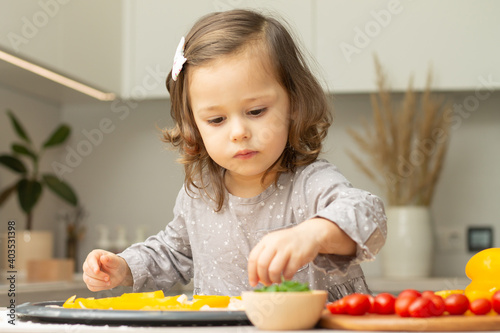 Cute little girl 2-4 in gray dress cooking pizza in kitchen. Kid arranges ingredients on pizza base