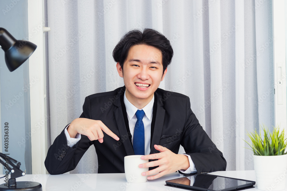 Work from home, Asian young businessman video conference call or facetime he looking to camera sitting on desk point coffee cup to invite colleagues drink coffee before event at home office Work from home, Asian young businessman video conference call or facetime he looking to camera sitting on desk point coffee cup to invite colleagues drink coffee before event at home office