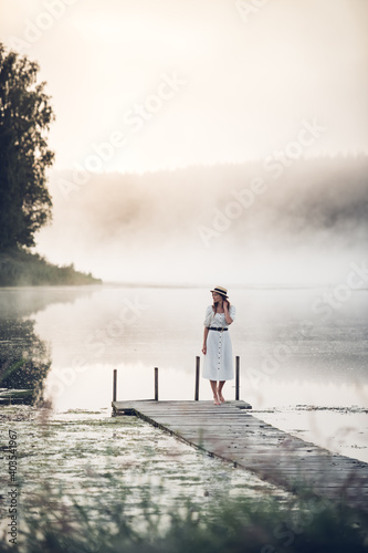 Woman in a white dress and hat standning on a wooden pier with a foggy lake at sunrise.