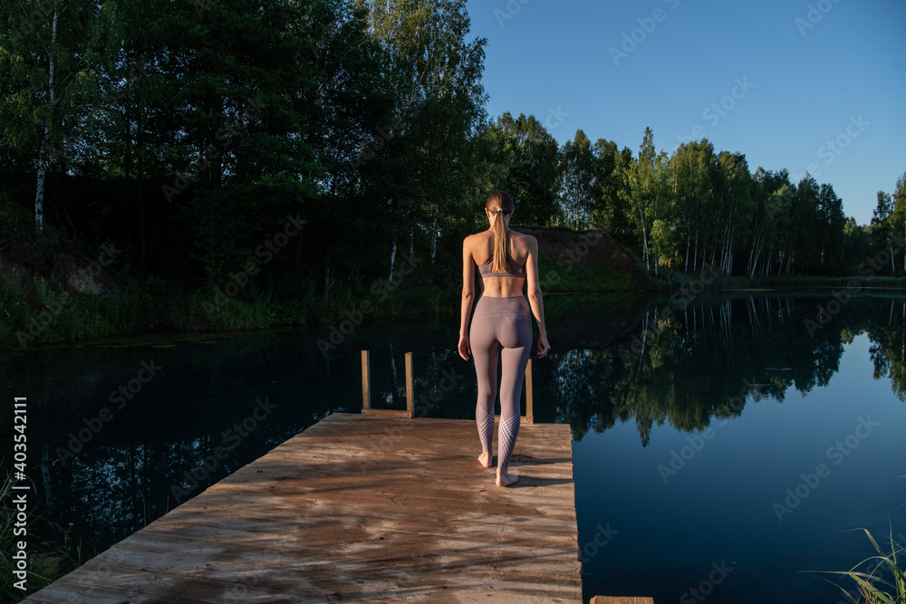 Young woman walks on wooden pier above forest lake scenery, preparing for morning yoga workout in nature. Young woman walks on wooden pier above forest lake scenery, preparing for morning yoga workout in nature.