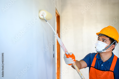 Young Asian painter Wear a protective mask to paint the indoor white walls with a paint roller in the new house.