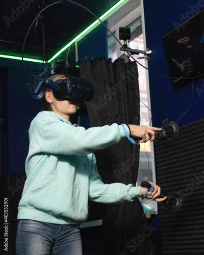 Young teenager girl using virtual reality headset with goggles and hands motion controllers in gaming zone.
