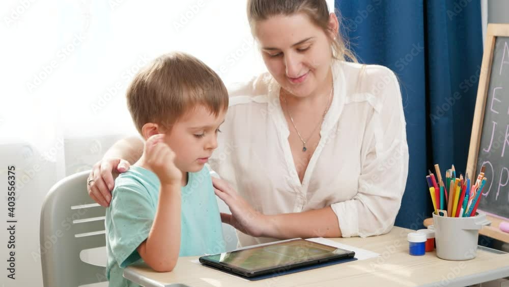 Smiling mother hugging and caressing her smart child studying on tablet computer. Little boy doing homework on digital tablet computer at home Smiling mother hugging and caressing her smart child studying on tablet computer. Little boy doing homework on digital tablet computer at home