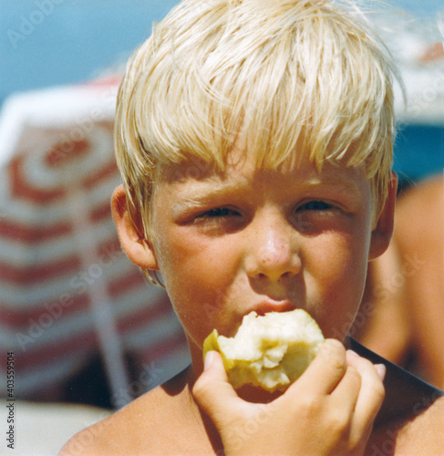 Vintage colorful retro 1979 image, close-up portrait of a young boy with blond hair and beach umbrella background eating an apple. photo