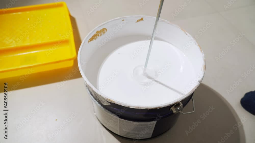 A worker mixes white paint in a bucket using an electric mixer. A worker mixes white paint in a bucket using an electric mixer.