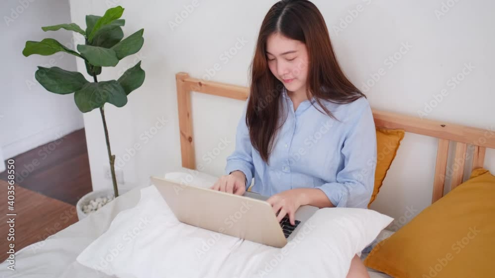 Asian young beautiful woman sit on bed and work with laptop in bedroom. Work at home with new normal lifestyle during Covid-19 pandemic in city concept.